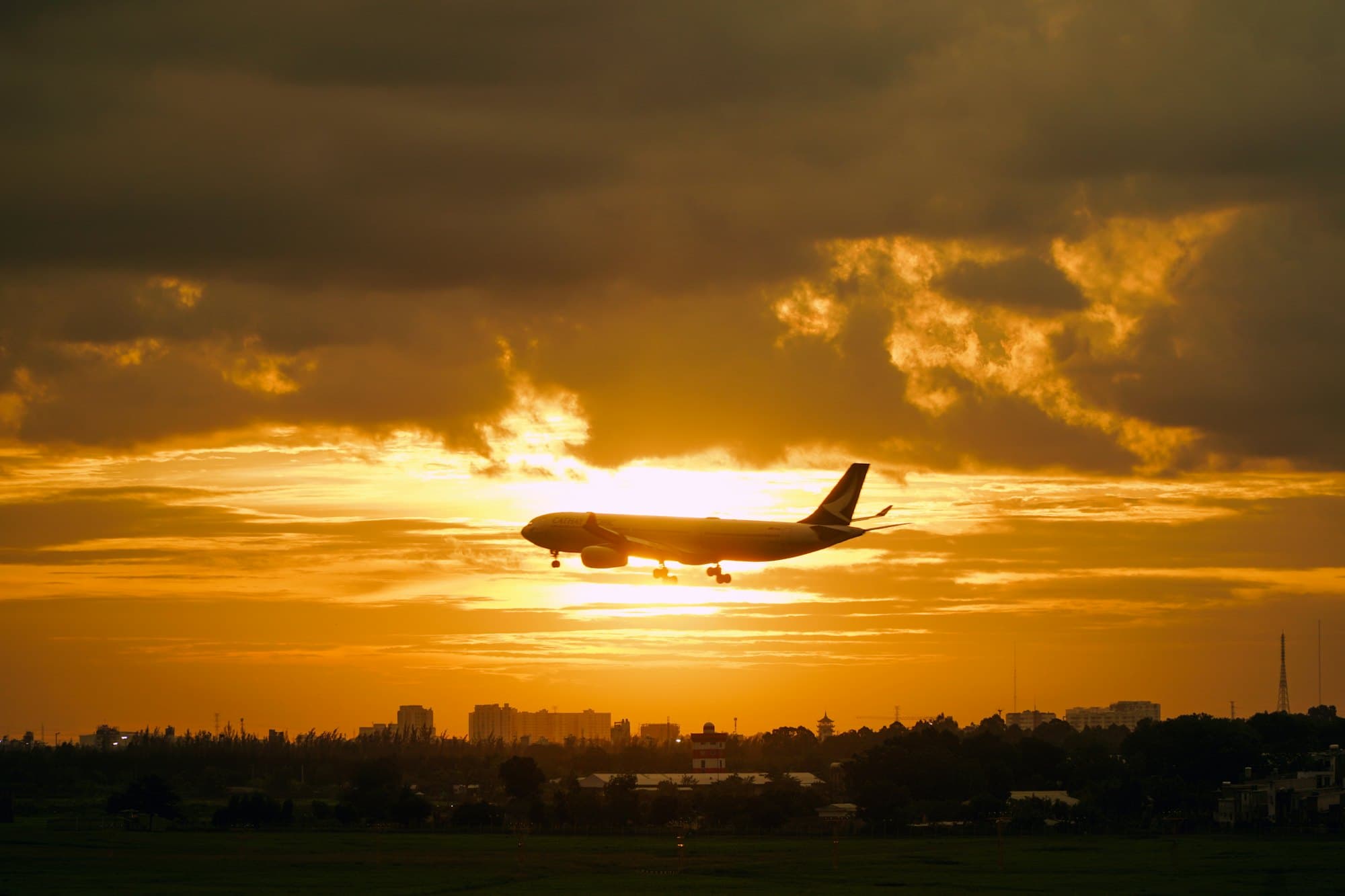 A passenger aircraft on final approach, silhouetted against a burning orange sunset above a tree line