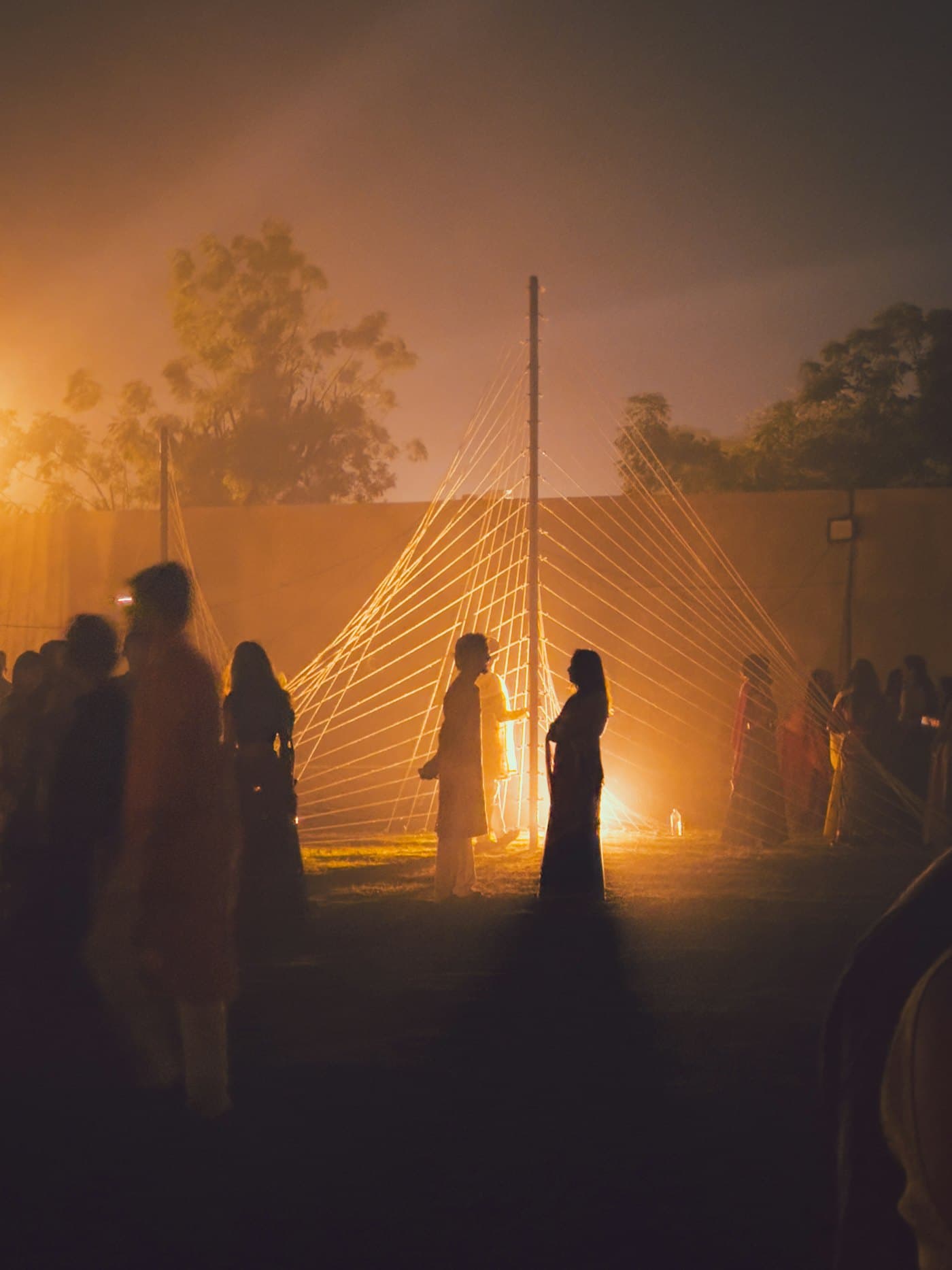 Wedding guests in silhouette on a lawn at night, warm light glowing through mist and a string installation