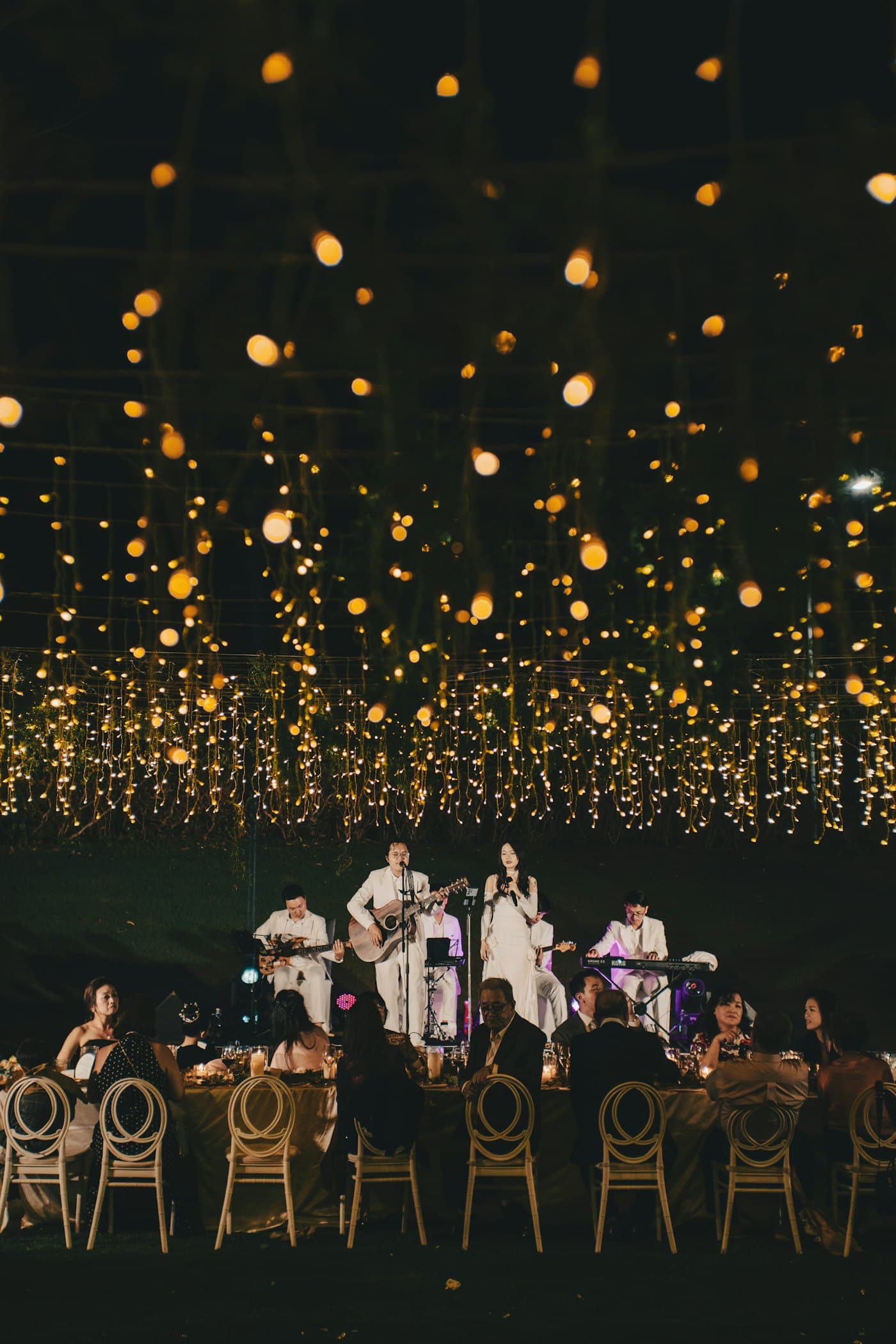 Guests dining at round tables beneath a dense canopy of hanging fairy lights at night