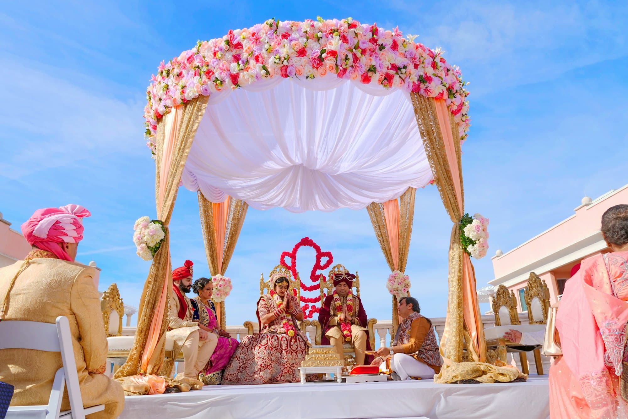 A bride and groom in traditional attire seated beneath a floral mandap canopy