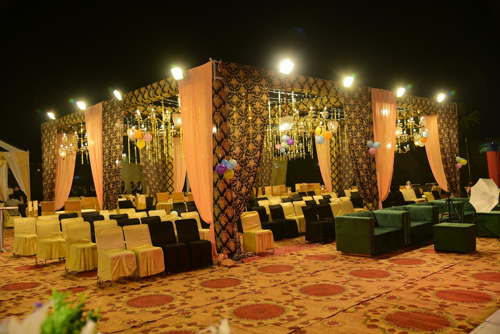 An open-air wedding pavilion at night, hung with chandeliers and draped in peach fabric, rows of chairs on a patterned carpet