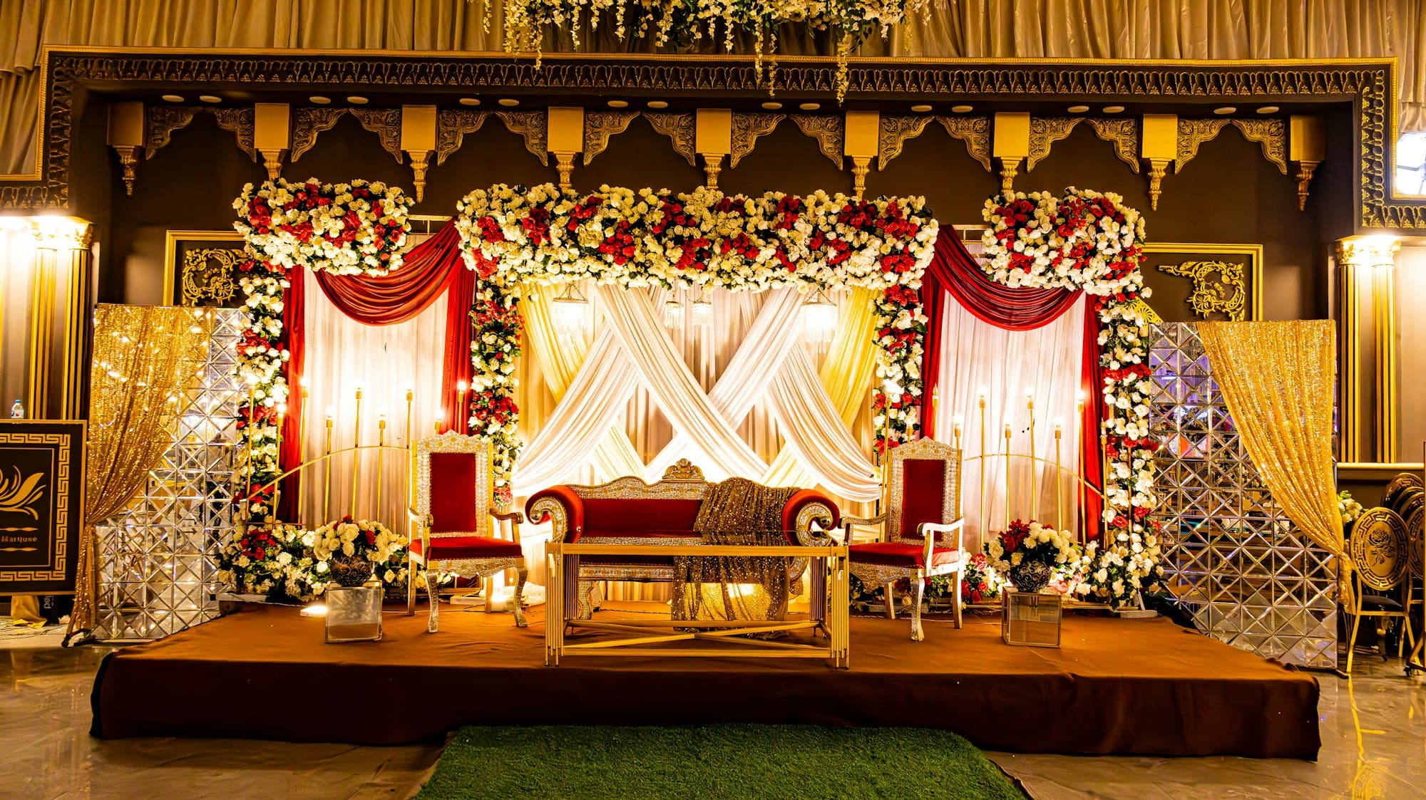 A decorated reception stage with floral backdrop, chandeliers and a couple's seating